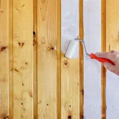 Ręka w rękawiczce maluje białą farbą drewniane panele ścienne. Tak, panele ścienne można malować, odnawiając ich wygląd.