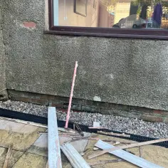 Na zewnątrz, przy ścianie domu, leżą resztki materiałów budowlanych, w tym fragmenty listwy przypodłogowej wodoodpornej.