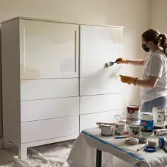 Kobieta w masce i rękawiczkach maluje białą szafę z płyty wiórowej. Na stole stoją puszki z farbą, pokazując, jak pomalować meble z płyty wiórowej.