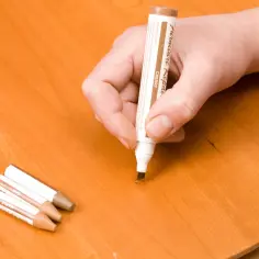 Dłoń trzyma marker do renowacji mebli, próbując zamaskować rysę na drewnianym stole. Obok leżą inne markery. To pokazuje, jak zmyć marker ze stołu, maskując uszkodzenia.