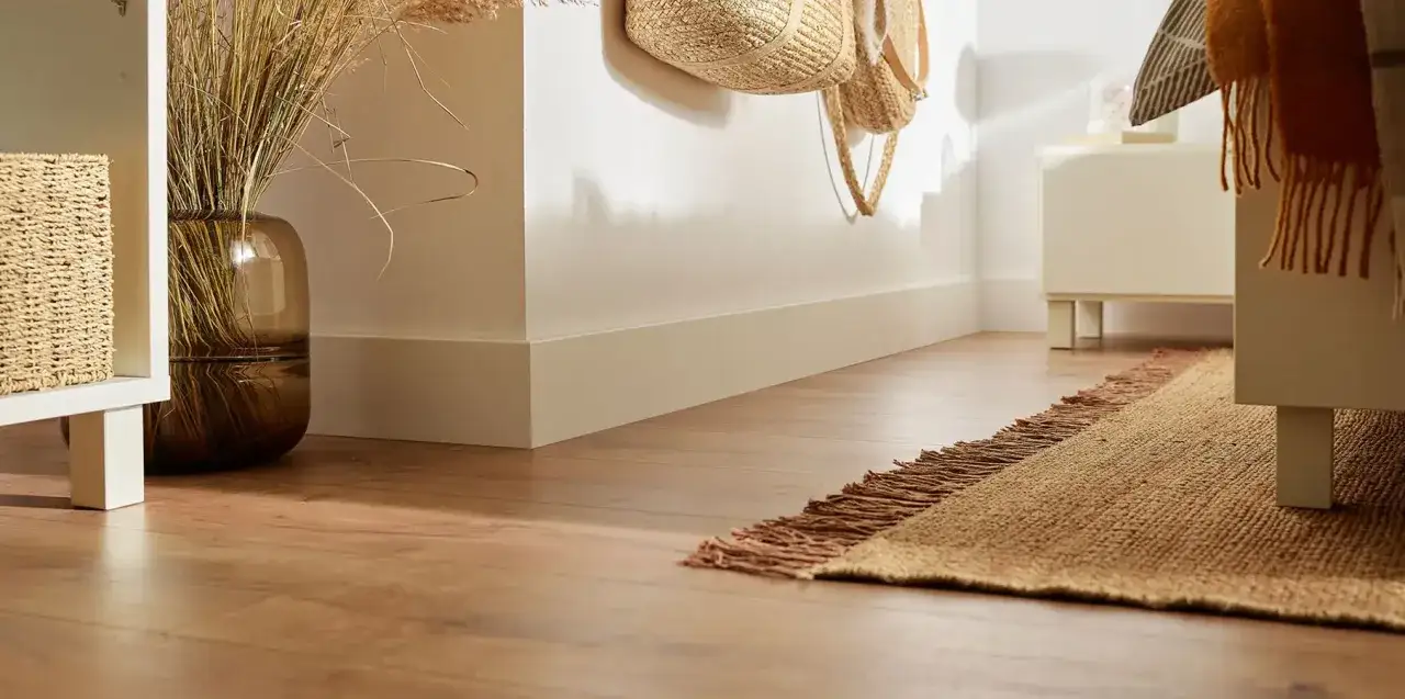 Elegancka listwa przypodłogowa złota subtelnie podkreśla drewnianą podłogę i jasne ściany. W tle wazon z trawami i plecione kosze.