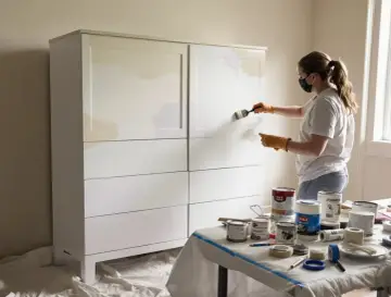Kobieta w masce i rękawiczkach maluje białą szafę z płyty wiórowej. Na stole stoją puszki z farbą, pokazując, jak pomalować meble z płyty wiórowej.