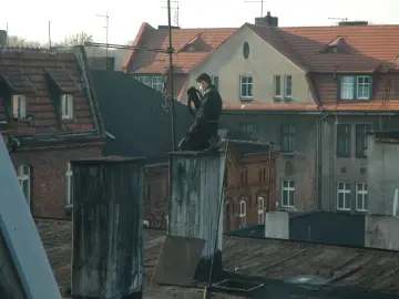 Kominiarz wykonuje przegląd kominiarski w mieszkaniu bez gazu, siedząc na dachu kamienicy.