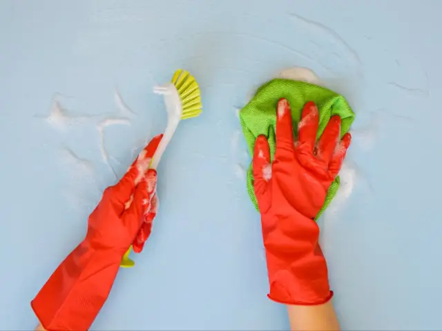 Ręce w czerwonych rękawiczkach czyszczą niebieską ścianę. Jedna ręka trzyma szczotkę, druga zieloną ściereczkę z pianą. Dowiedz się, jak zmyć wosk ze ściany.
