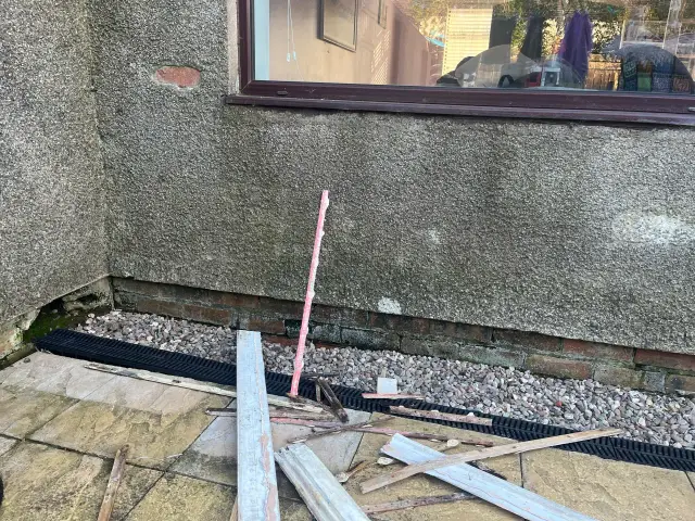 Na zewnątrz, przy ścianie domu, leżą resztki materiałów budowlanych, w tym fragmenty listwy przypodłogowej wodoodpornej.