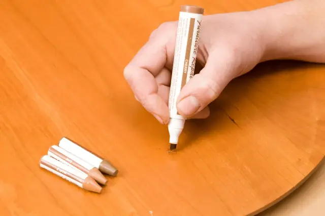 Dłoń trzyma marker do renowacji mebli, próbując zamaskować rysę na drewnianym stole. Obok leżą inne markery. To pokazuje, jak zmyć marker ze stołu, maskując uszkodzenia.